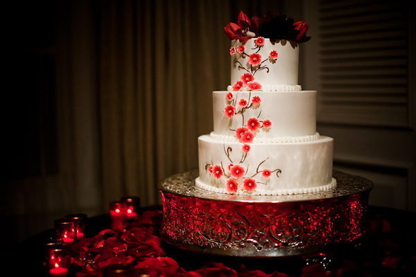 Beautiful white three tiered round wedding cake with coral and red floral accents on a vintage cake stand surrounded by red candles and red rose petals - wedding photo by Michael Norwood Photography
